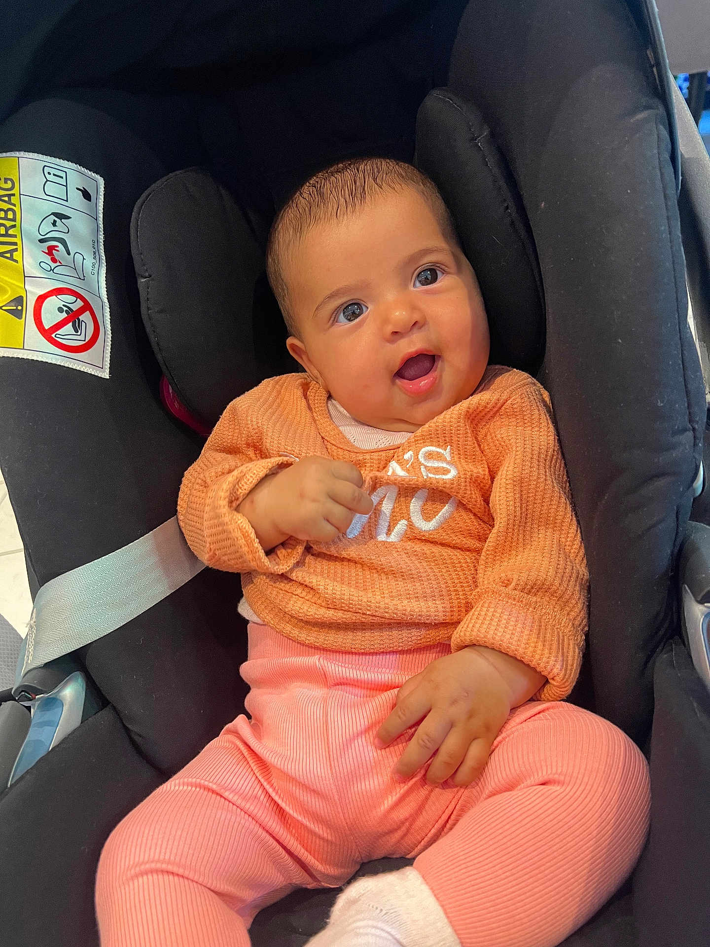 Nayla participe au concours pour gagner de l'argent avec cette photo : baby, child, infant, car_seat, orange_sweater, pink_pants, white_socks, smiling, happy, indoors, seatbelt, safety_label, person, cute, portrait, young_child, clothing, head, hand, face