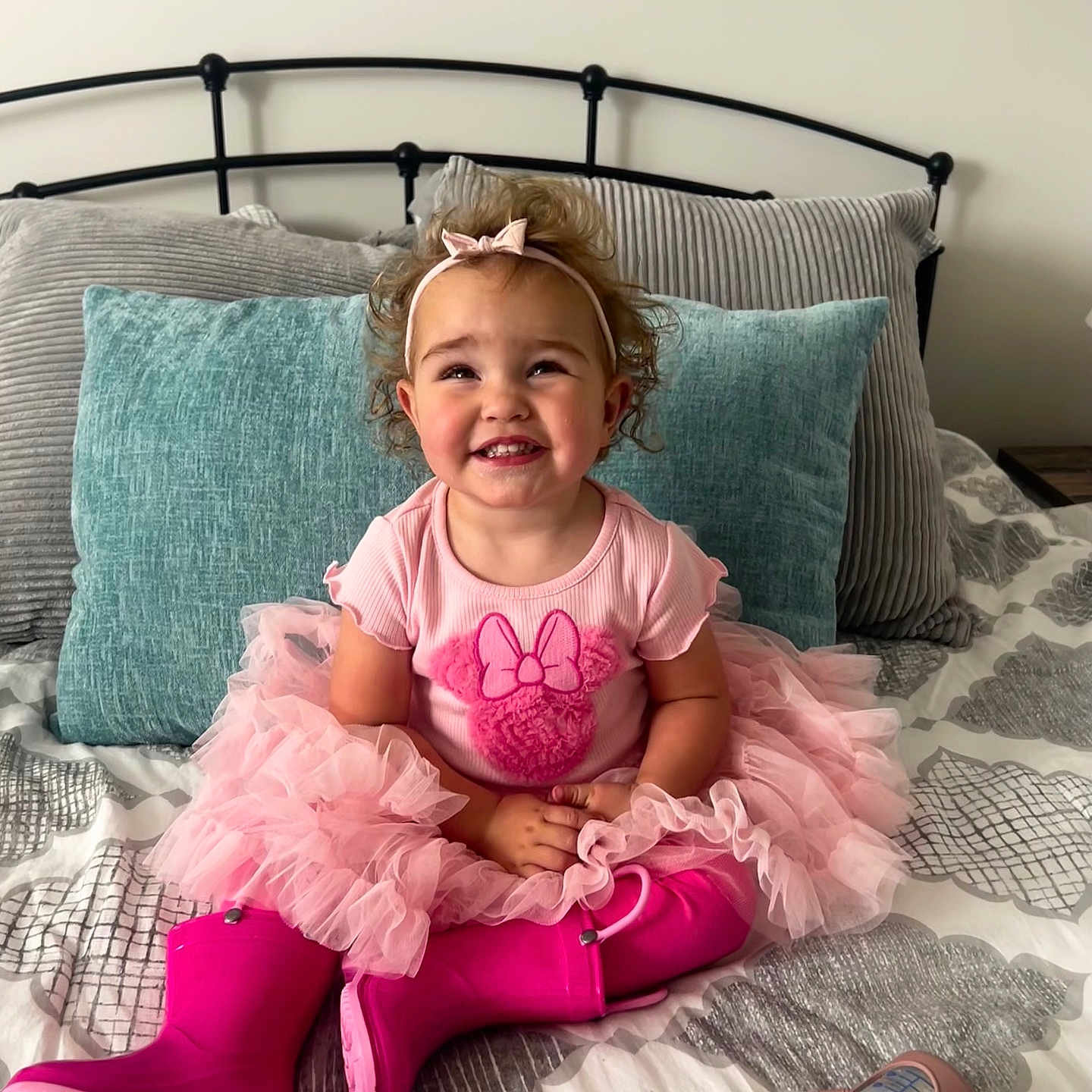 Elliana joined the competition — help win amazing prizes! toddler, child, pink_tutu, pink_boots, headband, smiling, bed, pillows, blanket, toy_bottle, indoor, curly_hair, happy, cute, sitting, clothing, person, face, home, playful