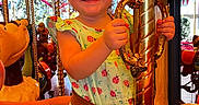 Elliana joined the competition — help win amazing prizes! child, girl, smile, carousel, amusement_park, ride, dress, ladybug_pattern, curly_hair, bow, hand, pole, colorful, fun, joy, happy, portrait, indoor, toy, person