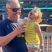 Elliana joined the competition — help win amazing prizes! child, toddler, man, ice_cream, yellow_shirt, sunglasses, stadium, outdoor, daylight, smiling, curly_hair, holding, happy, summer, food, family, fun, people, leisure, fence