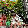 child, curly_hair, white_clothing, sandals, tree, red_flowers, green_leaves, stone_ledge, shipping_container, urban, outdoor, daylight, thoughtful, person, nature, road, car, sky, plant, rust