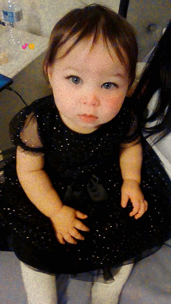 Isabella is registered to the contest to win money with this photo: toddler, child, baby, dress, black_dress, white_tights, blue_eyes, indoor, sitting, curious, person, face, hands, short_hair, floor, water_bottle, table, background, cute, portrait