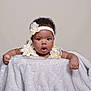 baby, infant, child, headband, flower, blanket, curly_hair, surprised_expression, soft_texture, cute, portrait, indoor, skin, hands, clothing, person, young_child, adorable, studio_photo, neutral_background