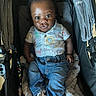 baby, child, stroller, jeans, shirt, animal_print, footwear, blanket, curly_hair, indoors, seated, person, face, wide_eyes, cute, casual_clothing, infant, baby_shoes, comfort, portrait