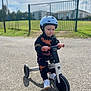 balance_bike, bike, casual_wear, child, clothing, daytime, fence, greenery, helmet, outdoor, pavement, person, play, serious_expression, shadow, shoes, sunny, toddler, toy, young_child