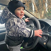 Thibaut participe au concours pour gagner de l'argent avec cette photo : toddler, child, car_interior, steering_wheel, winter_clothing, hat, jacket, gray_pants, curious, blue_eyes, hand_on_wheel, car_seat, dashboard, window, trees, daylight, indoors, seat_belt, cute, playful
