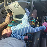Layton is registered to the contest to win money with this photo: baby, infant, newborn, car_interior, steering_wheel, tattooed_arm, hand, lap, striped_onesie, onesie, sunlight, shadow, center_console, cup_holder, water_bottle, clutter, seat, door_panel, fabric_pattern, skin