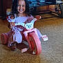 child, smile, tricycle, pink_tricycle, curly_hair, purple_dress, indoor, dvd_stack, shelf, tile_floor, playing, happy, toy, pedal, shoe, portrait, furniture, wood_paneling, hands_on_handlebars, cute
