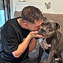 affection, affectionate, black_tshirt, bond, casual_clothing, comfort, couch, dog, face, gray_dog, home, human_dog_interaction, indoor, living_room, love, man, pet, seated, shorts, white_chest_marking