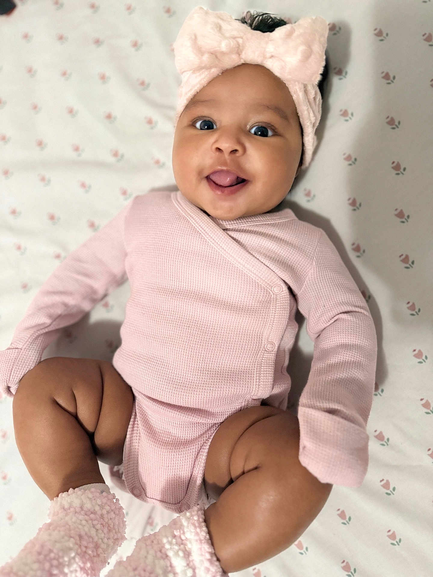 Ivonne joined the competition — help win amazing prizes! baby, infant, pink_clothing, headband, smiling, tongue_out, cute, lying_down, blanket, pattern, floral, soft_texture, cozy, happy, adorable, child, skin, face, legs, footwear