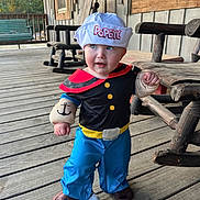 Brady is registered to the contest to win money with this photo: toddler, child, costume, popeye, sailor_hat, plush_gloves, blue_eyes, wooden_porch, rocking_chair, outdoor, daylight, curious_expression, blue_pants, black_shirt, red_collar, footwear, wooden_floor, person, portrait, cute
