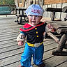 toddler, child, costume, popeye, sailor_hat, plush_gloves, blue_eyes, wooden_porch, rocking_chair, outdoor, daylight, curious_expression, blue_pants, black_shirt, red_collar, footwear, wooden_floor, person, portrait, cute