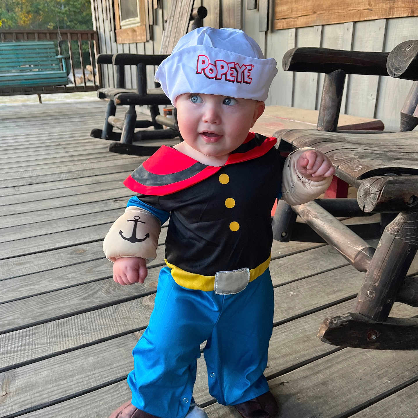 Brady is registered to the contest to win money with this photo: toddler, child, costume, popeye, sailor_hat, plush_gloves, blue_eyes, wooden_porch, rocking_chair, outdoor, daylight, curious_expression, blue_pants, black_shirt, red_collar, footwear, wooden_floor, person, portrait, cute