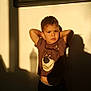 child, boy, portrait, standing, shadow, wall, sunlight, golden_hour, expression, t_shirt, bear_graphic, cartoon, indoor, window_frame, fan, silhouette, pose, casual_clothing, home_interior, dramatic_lighting