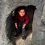 child, toddler, boy, face, red_sweater, jeans, sneakers, crouching, rock, cave, playground, peekaboo, expression, hands, portrait, adventure, curious, smile, clothing, shoe