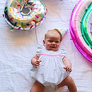 Rose a rejoint le concours — aidez-le/la à gagner de superbes lots ! baby, child, party_hat, white_dress, balloon, donut_balloon, rainbow_balloon, celebration, smiling, happy, infant, cute, lying_down, indoor, colorful, birthday, fun, joyful, playful, festive
