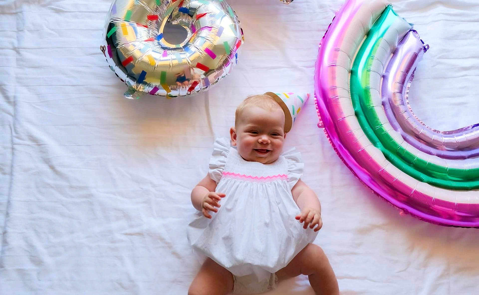 Rose a rejoint le concours — aidez-le/la à gagner de superbes lots ! baby, child, party_hat, white_dress, balloon, donut_balloon, rainbow_balloon, celebration, smiling, happy, infant, cute, lying_down, indoor, colorful, birthday, fun, joyful, playful, festive