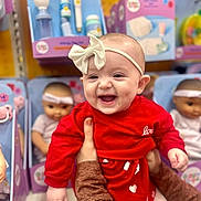Lyra is registered to the contest to win money with this photo: baby, smiling, red_clothing, headband, bow, toy_store, dolls, shelf, happy, infant, person, hands, cute, child, indoor, retail, shopping, face, joy, playful