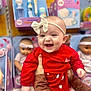baby, smiling, red_clothing, headband, bow, toy_store, dolls, shelf, happy, infant, person, hands, cute, child, indoor, retail, shopping, face, joy, playful