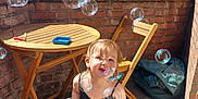 Ylliana a rejoint le concours — aidez-le/la à gagner de superbes lots ! balcony, black_dress, brick_wall, bubbles, casual, child, daytime, feet, greenery, happy, outdoor, person, playful, shadow, smiling, sunlight, toddler, window, wooden_chair, wooden_table