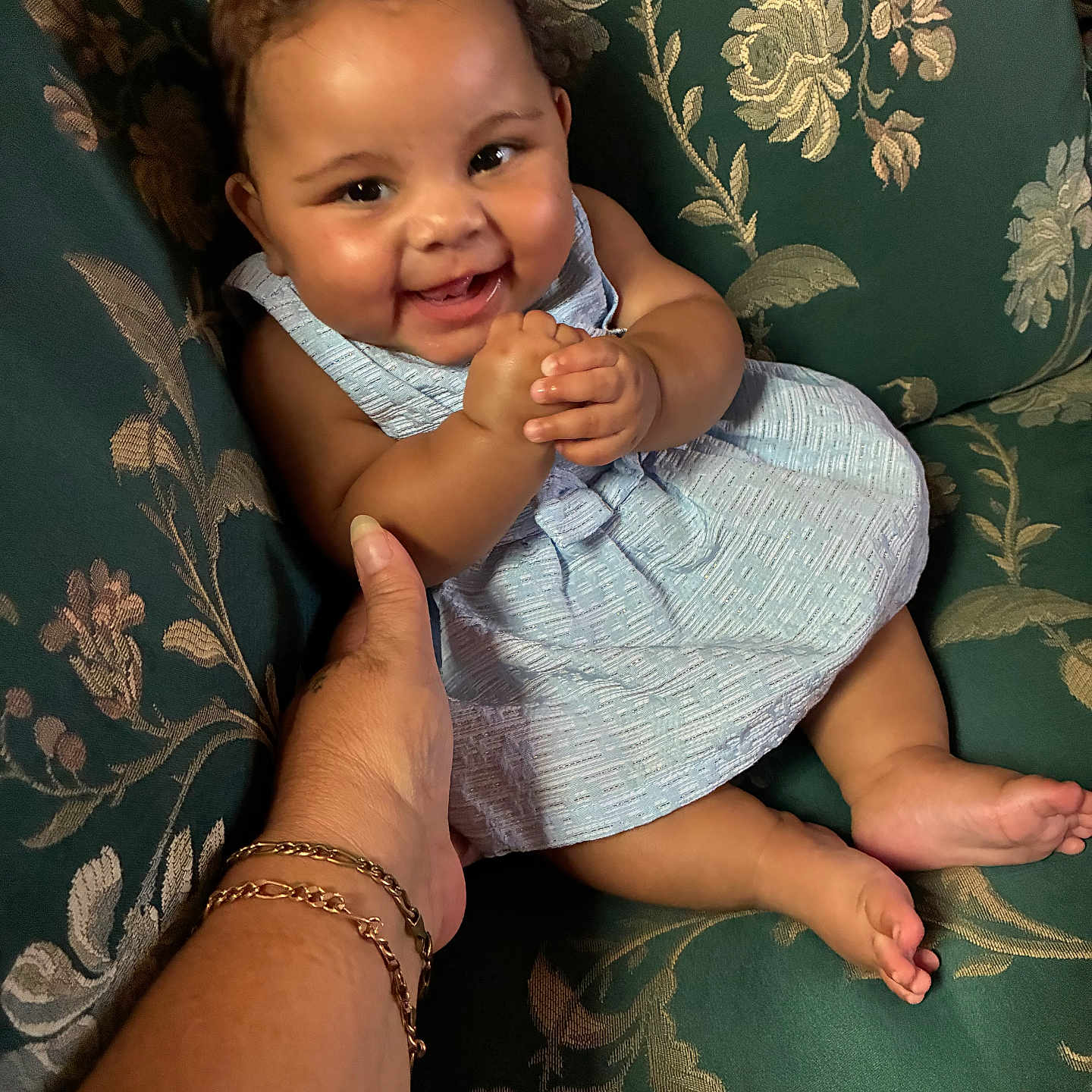 Jalayah is registered to the contest to win money with this photo: baby, infant, child, smiling, happy, portrait, seated, armchair, floral_pattern, blue_dress, hands, adult_hand, bracelet, indoor, furniture, close_up, bare_feet, joyful, family, candid