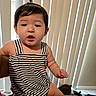 baby, child, indoor, striped_clothing, romper, curly_hair, pony_tail, face, person, sitting, vertical_blinds, carpet, window, natural_light, expression, cute, infant, hand, foot, room