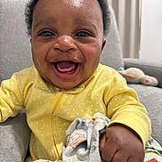 Prince is registered to the contest to win money with this photo: baby, child, smiling, yellow_onesie, blanket, couch, indoor, happy, face, infant, curly_hair, plush_toy, furniture, portrait, cute, hand, clothing, home, seated, toddler