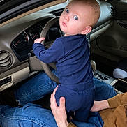 Weston is registered to the contest to win money with this photo: baby, child, car, steering_wheel, blue_clothing, adult, jeans, brown_shirt, interior, vehicle, seat, dashboard, curious, grip, hands, person, window, nature, forest, candid