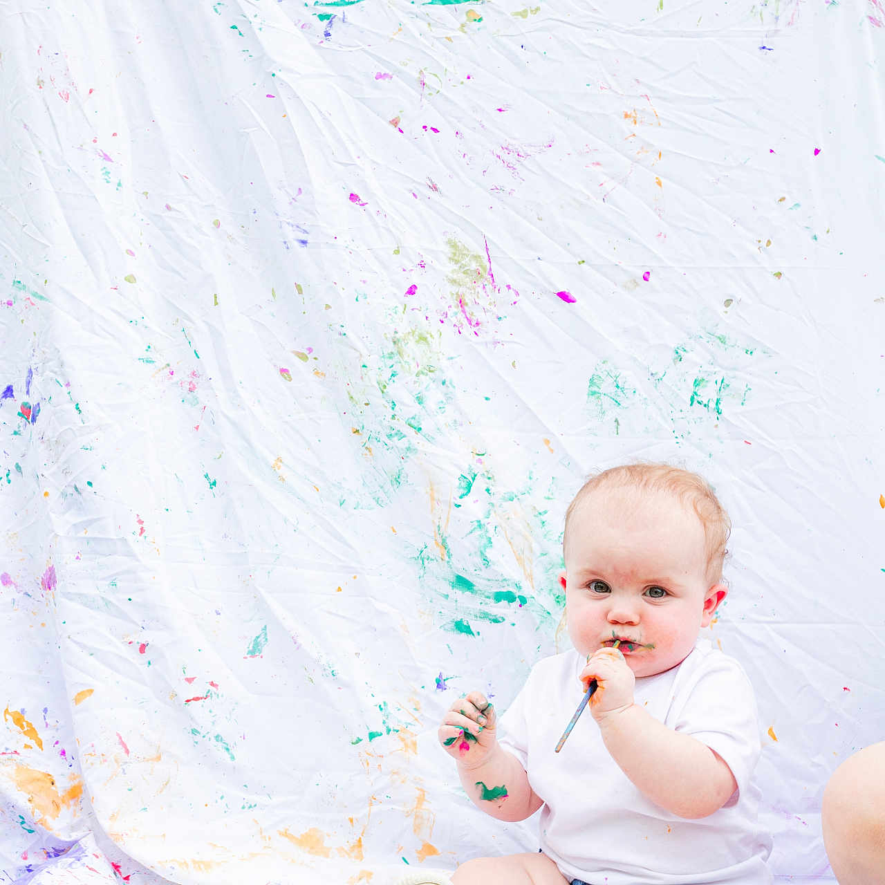 Lily is registered to the contest to win money with this photo: art, baby, casual_clothing, child, colorful, creative, curious, face_paint, grass, hand_paint, messy, outdoor, paint, paintbrush, playful, portrait, shorts, sitting, white_sheet, white_shirt