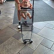 Aitana joined the competition — help win amazing prizes! baby, child, smiling, shopping_cart, store_entrance, indoor, adult, parent, headband, pink_bow, seated, happy, portrait, floor_tiles, doors, retail, family, cart, shopping, playful