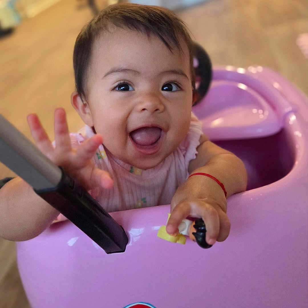 Aitana is registered to the contest to win money with this photo: baby, child, infant, smiling, happy, portrait, indoor, playroom, ride_on_toy, pink, toy_vehicle, sitting, reaching, hand, open_mouth, brown_hair, face, family, home, number_three