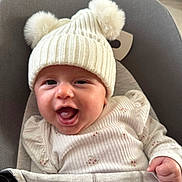 Esme joined the competition — help win amazing prizes! baby, smiling, hat, pom_poms, white_clothing, baby_chair, indoor, cute, happy, infant, face, person, child, cozy, seat, knit, puffed_sleeves, closeup, soft_lighting, portrait