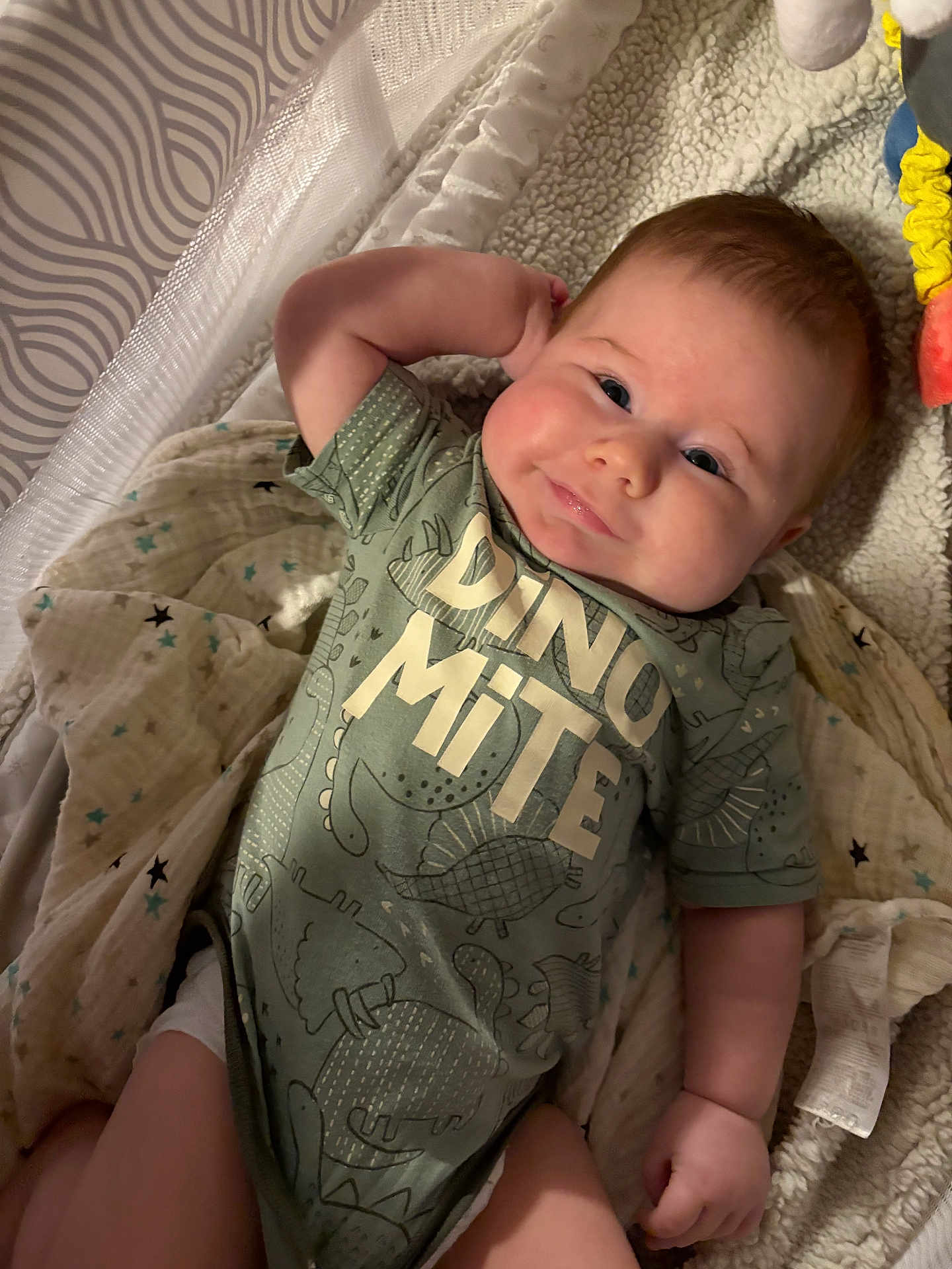 Lucious is registered to the contest to win money with this photo: baby, infant, onesie, dinosaur, blanket, smile, face, arm, hand, fabric, soft, cute, lying_down, child, relaxed, indoors, cozy, text_on_clothing, cheeks, head