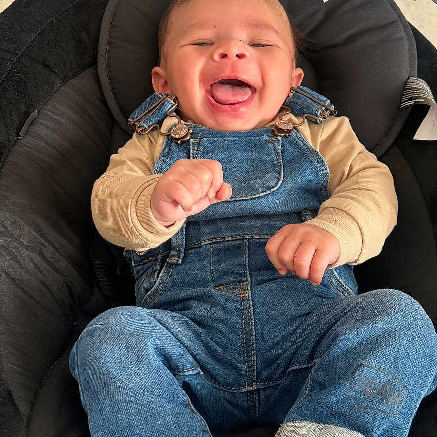 Nahyl participe au concours pour gagner de l'argent avec cette photo : baby, beige_shirt, black_cushion, candid, child, clothing, comfortable, cute, denim_overalls, face, feet, hands, happy, indoors, infant, joyful, portrait, sitting, smiling, young
