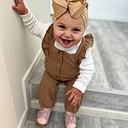 Lyana a rejoint le concours — aidez-le/la à gagner de superbes lots ! toddler, child, smiling, headband, bow, brown_clothing, white_shirt, pink_shoes, indoor, floor, steps, hand, face, happy, cute, person, portrait, holding_wall, young_child, casual_wear