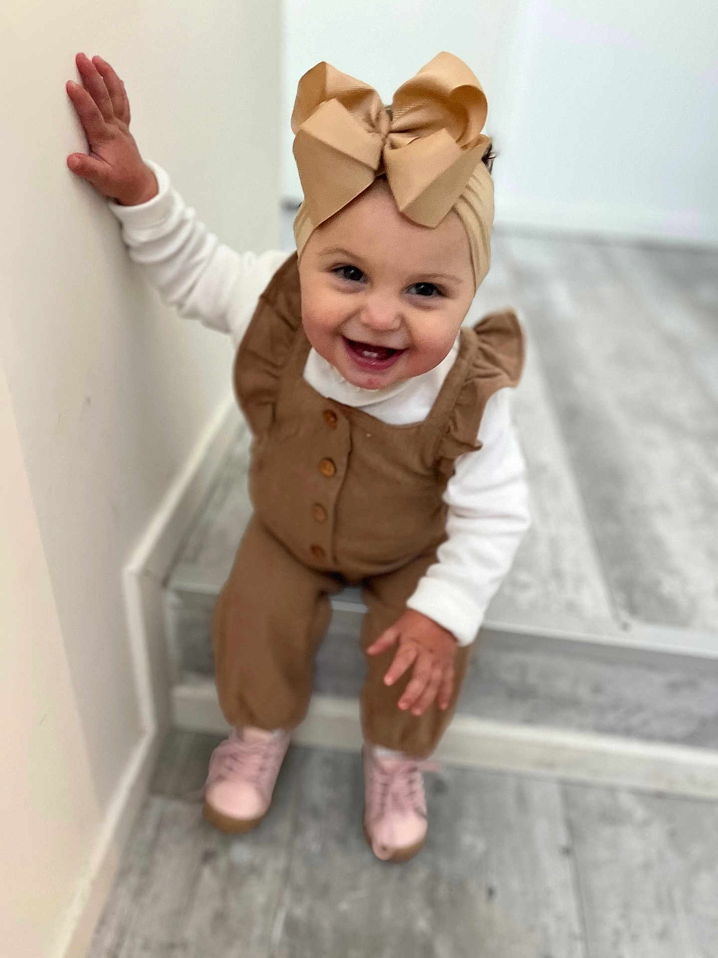Lyana a rejoint le concours — aidez-le/la à gagner de superbes lots ! toddler, child, smiling, headband, bow, brown_clothing, white_shirt, pink_shoes, indoor, floor, steps, hand, face, happy, cute, person, portrait, holding_wall, young_child, casual_wear