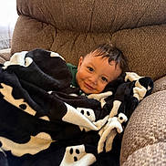 Mateo is registered to the contest to win money with this photo: baby, child, blanket, ghost_pattern, brown_armchair, smile, cozy, indoor, cute, snuggled, comfort, face, person, soft, plush, happy, young_child, hair, relaxing, portrait