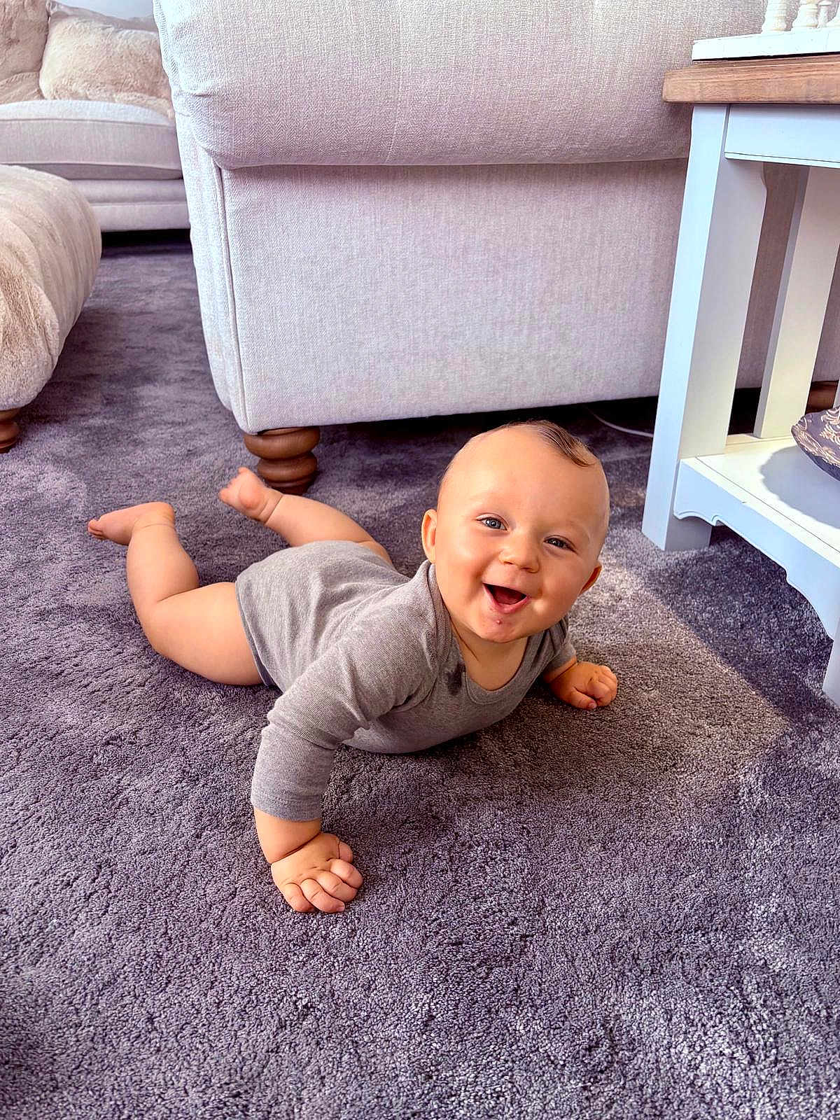 Teddie is registered to the contest to win money with this photo: baby, infant, smiling, carpet, living_room, furniture, sofa, table, floor, child, cute, happy, indoors, person, skin, clothing, head, eyes, hand, legs