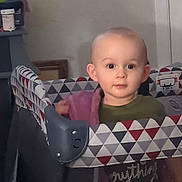 Spencer is registered to the contest to win money with this photo: baby, blanket, child, curious, door, expression, face, furniture, geometric_pattern, green_shirt, head, home, indoor, person, playpen, purple, room, standing, toddler, young_child