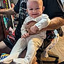 adult, baby, chair, child, clothing, face, floor, hand, happy, holding, household_items, indoor, pajamas, person, shelf, shorts, sitting, smiling, tshirt, wood_floor