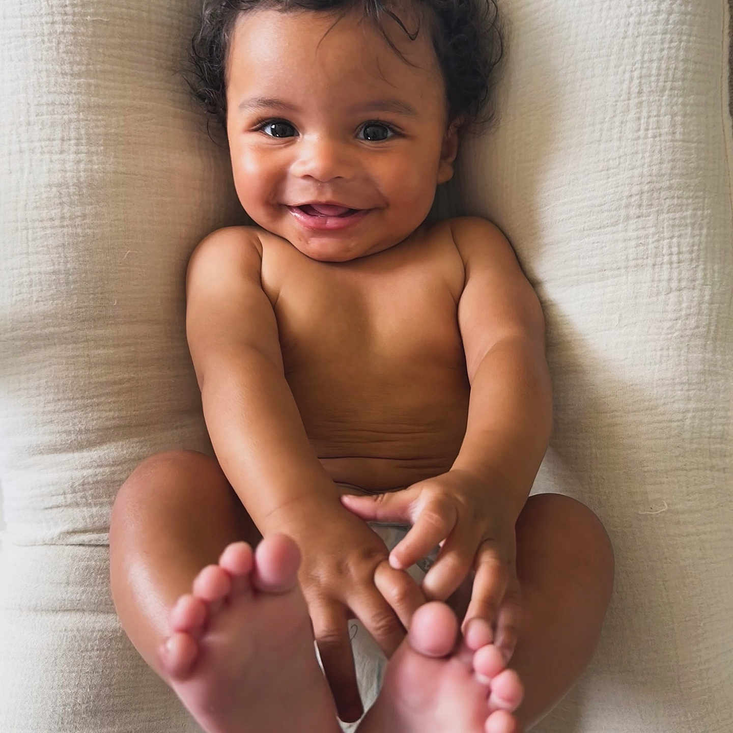 Devin is registered to the contest to win money with this photo: baby, child, smile, curly_hair, bare_skin, feet, hands, cushion, white_background, infant, happy, portrait, cute, playful, closeup, lying_down, human, skin, indoors, person