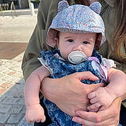 évy a rejoint le concours — aidez-le/la à gagner de superbes lots ! baby, infant, pacifier, hat, floral_hat, holding_hands, adult_hands, denim_outfit, outdoor, sunlight, cobblestone, street, woman, child, cute, portrait, casual_clothing, daytime, person, closeup