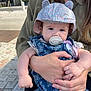 baby, infant, pacifier, hat, floral_hat, holding_hands, adult_hands, denim_outfit, outdoor, sunlight, cobblestone, street, woman, child, cute, portrait, casual_clothing, daytime, person, closeup