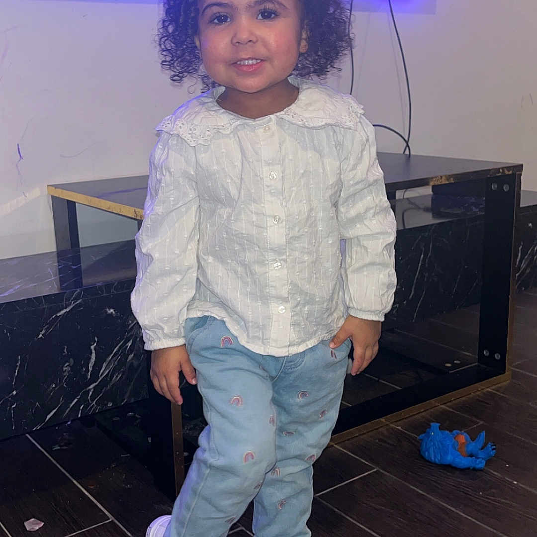 Saïana participe au concours pour gagner de l'argent avec cette photo : black_marble_table, blue_pants, casual_clothing, child, curly_hair, cute, floor, happy, indoor, modern_interior, person, purple_light, rainbow_pattern, smiling, standing, toddler, toy, white_blouse, white_sneakers, wooden_floor