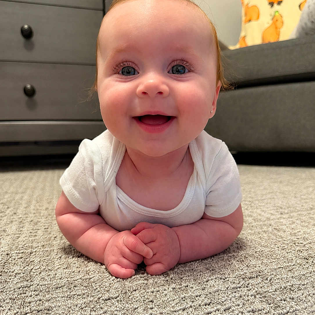 Lilith is registered to the contest to win money with this photo: baby, smiling, carpet, indoor, crawling, happy, child, furniture, drawer, pillow, couch, white_clothing, face, person, cute, infant, baby_girl, floor, room, home