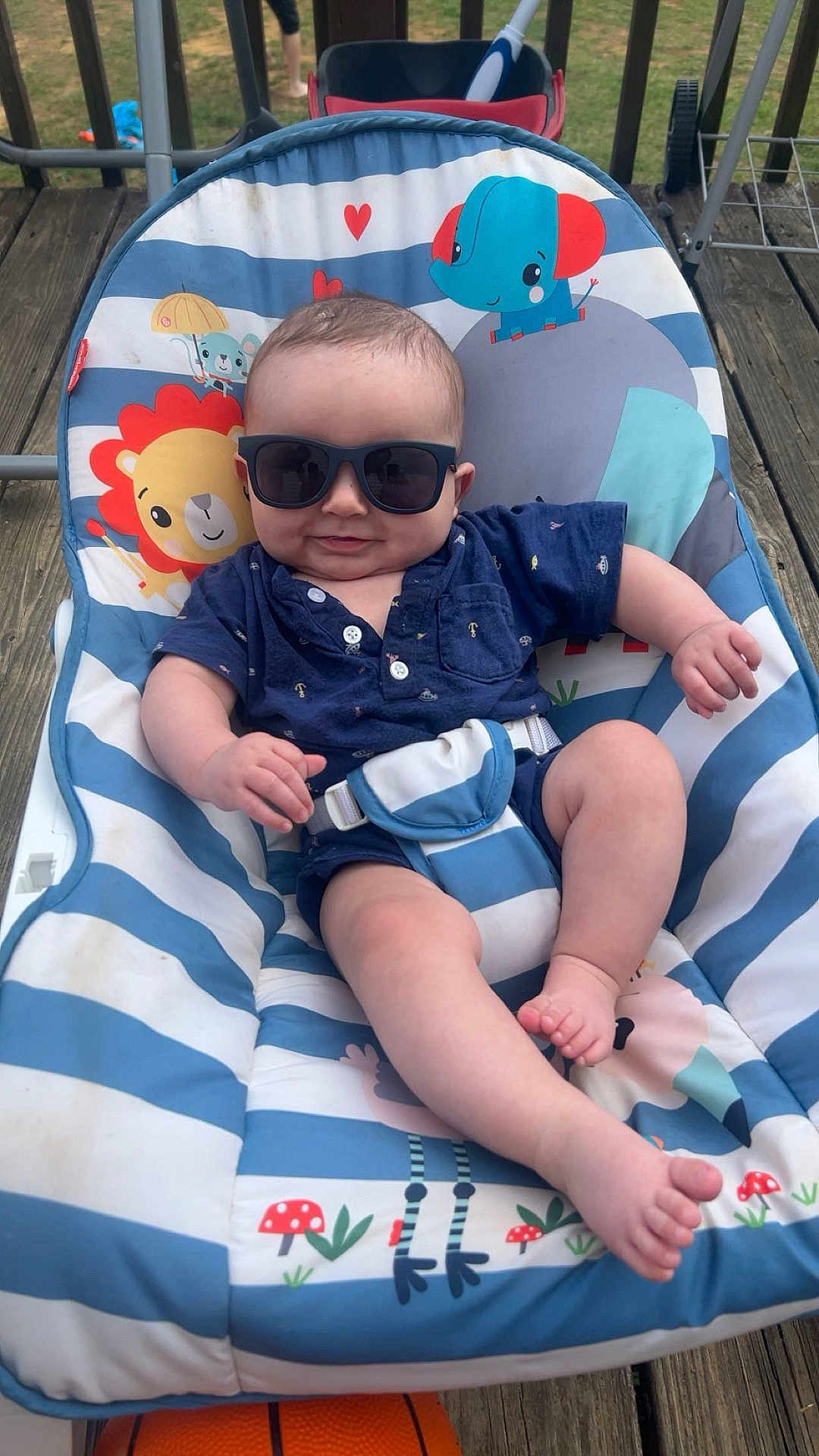 Logan is registered to the contest to win money with this photo: baby, sunglasses, blue_clothing, striped_baby_seat, lion_print, elephant_print, outdoor, wooden_deck, basketball, infant, smiling, relaxed, summer, child, playful, seat_belt, cute, sunny, toy, baby_chair
