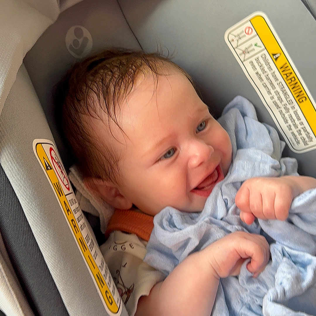 Xander is registered to the contest to win money with this photo: baby, car_seat, blanket, smiling, infant, child, blue_blanket, happy, face, head, clothing, seatbelt, padding, warning_label, person, cute, newborn, indoors, portrait, comfort