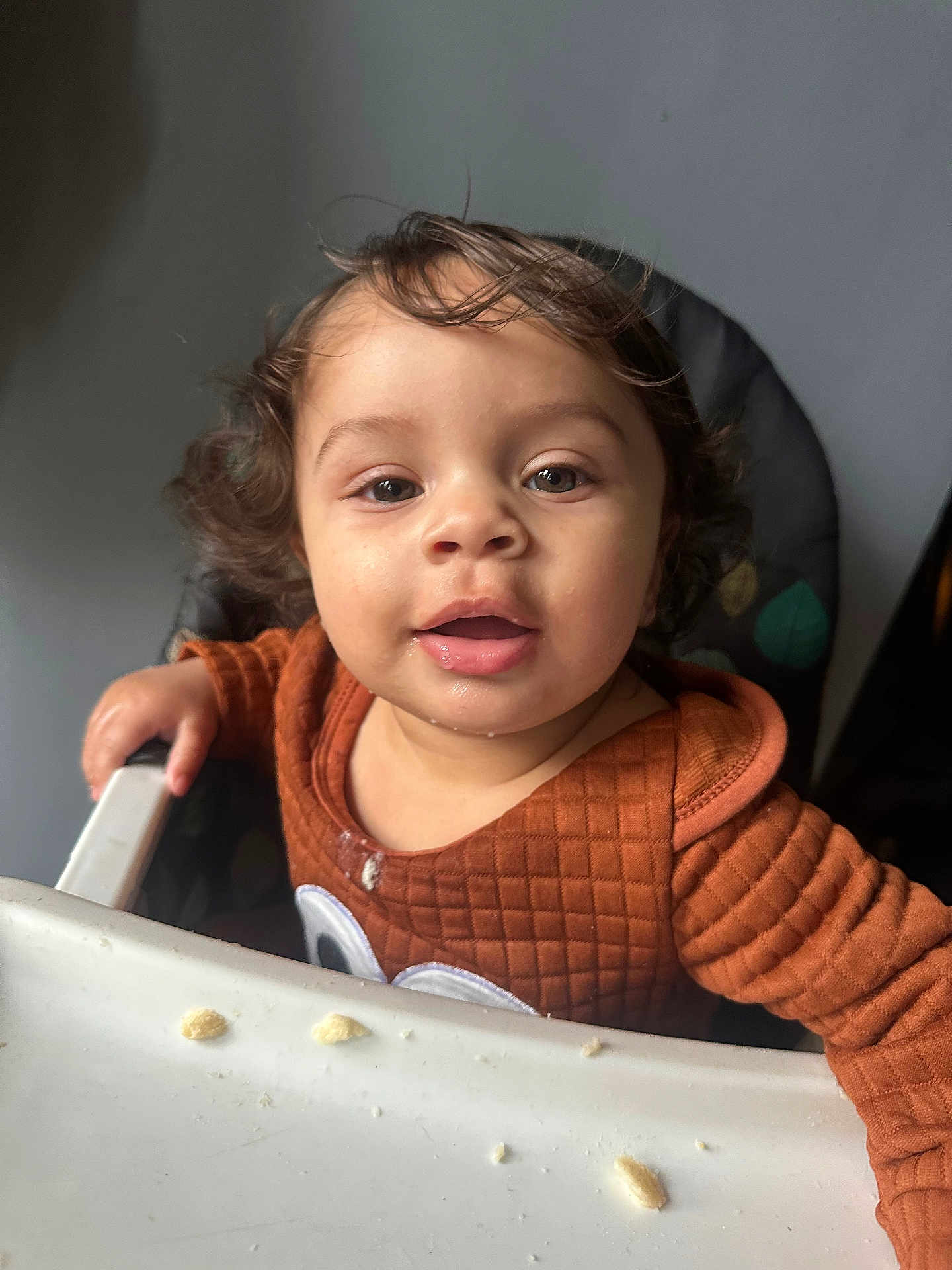 Macai is registered to the contest to win money with this photo: toddler, child, high_chair, orange_clothing, curly_hair, crumbs, indoor, face, portrait, baby_food, cute, smiling, young_child, feeding_time, happy, person, seat, curious, close_up, expression