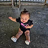 baby, child, toddler, sitting, smiling, face, curly_hair, ponytail, black_shirt, dinosaur_shirt, pink, sneakers, shoes, outdoor, pavilion, wooden_railings, concrete_floor, chalk_marks, sunlight, portrait