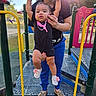 baby, child, adult, playground, playground_equipment, swing_set, hanging, smiling, outdoor, park, sunlight, portrait, parent, support, infant, baby_shoes, black_clothing, colorful_equipment, grass, family